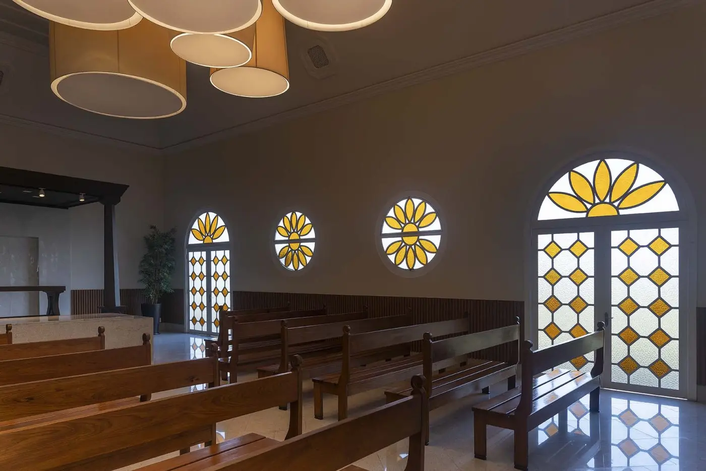 Interior da capela do Parque dos Ipês: bancos de madeira enfileirados, vitrais amarelos em desenho de flor e luz natural entrando pelas janelas em arco.