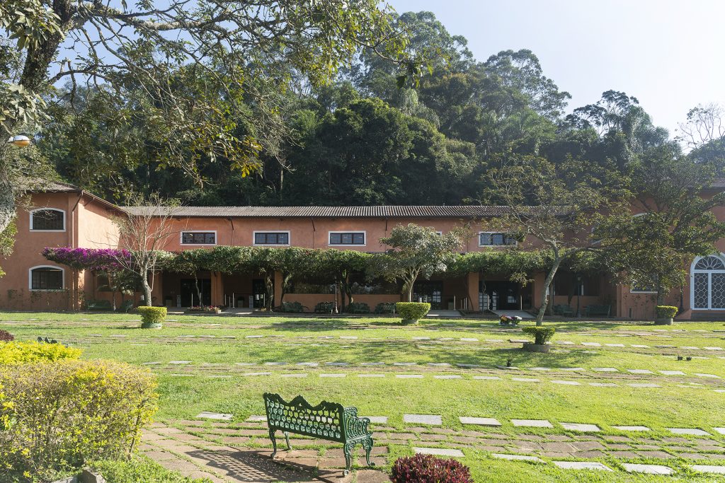 Jardim do Parque dos Ipês, com edificação em terracota, buganvília, lápides rasas no gramado e banco de ferro à sombra da mata.