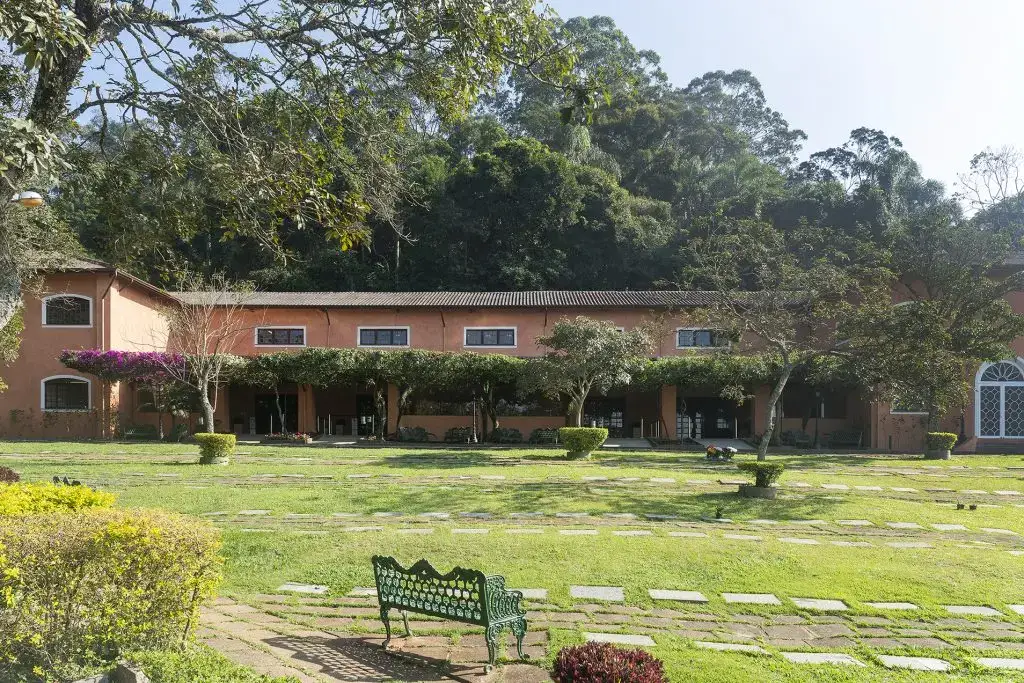 Jardim do Parque dos Ipês, com edificação em terracota, buganvília, lápides rasas no gramado e banco de ferro à sombra da mata.