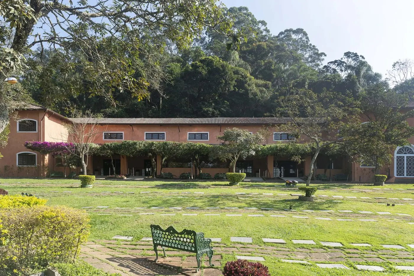 Gramado do Parque dos Ipês com placas rasas, banco de ferro verde em primeiro plano, edificação em terracota com buganvília e a mata ao fundo.